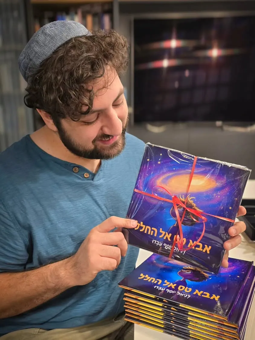 דניאל מחייך ומחזיק עותקים של הספר, עטופים בסרט אדום.